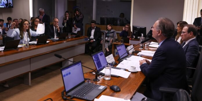 Woman speaker at a conference table raises a document while addressing a formal meeting, surrounded by colleagues with laptops and notes.