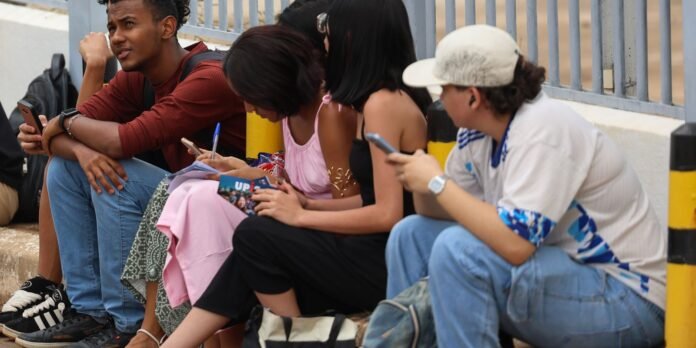 Group of diverse young people sitting along a curb near a railing, all absorbed in their phones or notes.