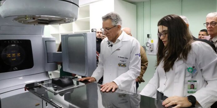 Medical team in white coats stands beside a radiotherapy linear accelerator (Clinac) with a patient immobilization mask on the treatment table, observing the setup.