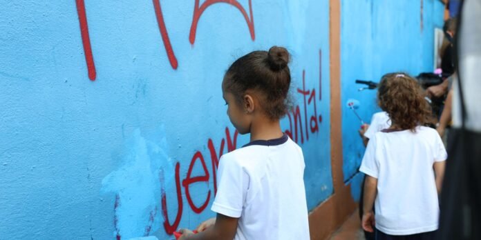 Two young girls in white shirts painting a blue wall mural with red letters.