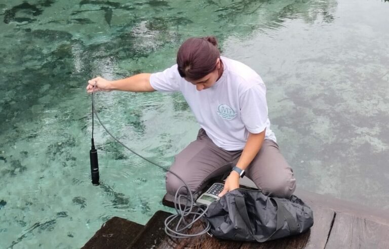 Researcher lowering a water quality probe into turquoise lake water, with a handheld meter and gear bag on the dock.