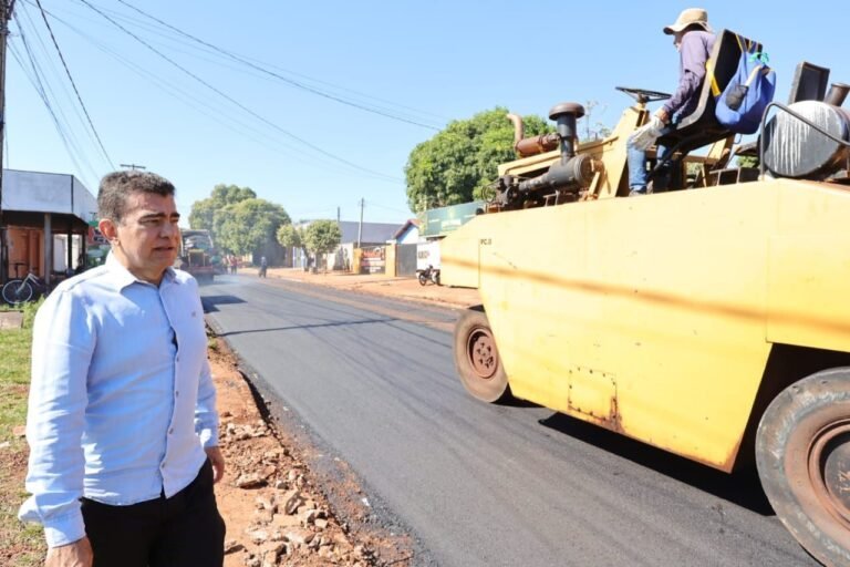 A man in a light blue shirt watches a yellow road roller paving a street.