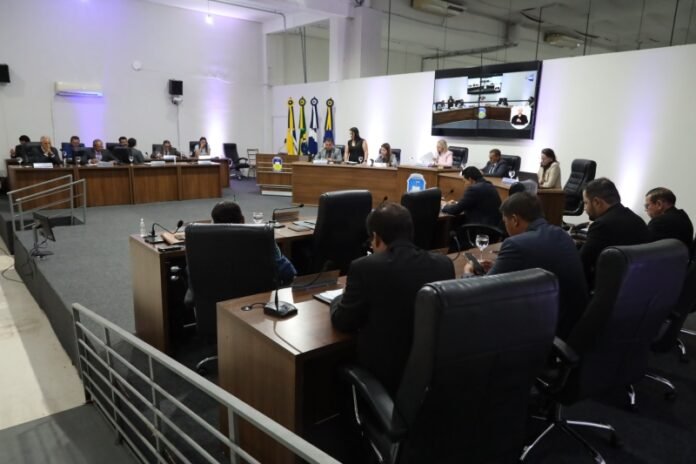 City council meeting in a modern chamber with officials at curved desks, microphones, flags behind, and a large screen on the wall.