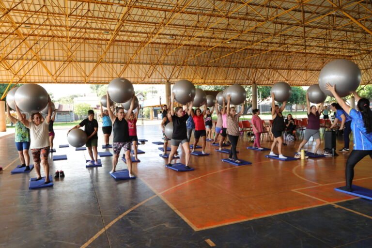 Group fitness class under a yellow-roofed pavilion, participants lift large gray stability balls above their heads on blue mats.
