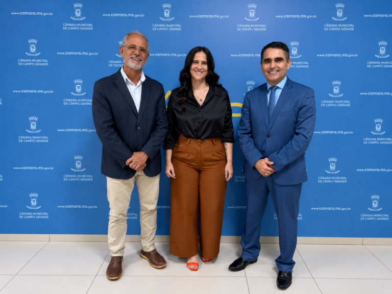 Three people pose in front of a blue backdrop with government logos: left man in navy blazer and beige pants, middle woman in black top and brown trousers, right man in blue suit.
