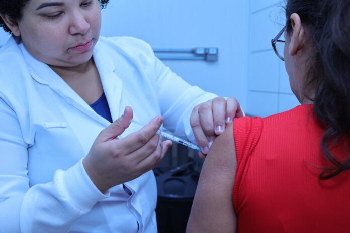 Healthcare professional administers a vaccination to a patient's upper arm with a syringe and needle visible, in a clinical setting.