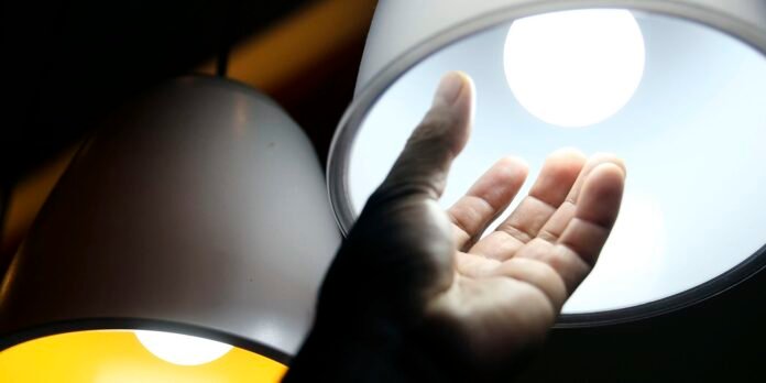 A hand reaching toward a bright circular lamp shade, with light washing over the palm in a dim room.