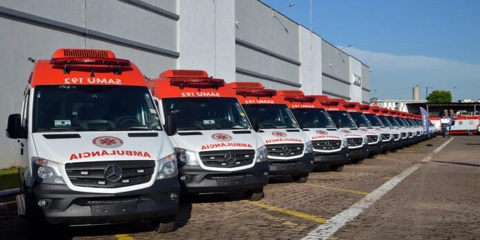 Line of white ambulances with red tops and emergency lights parked along a gray building complex.
