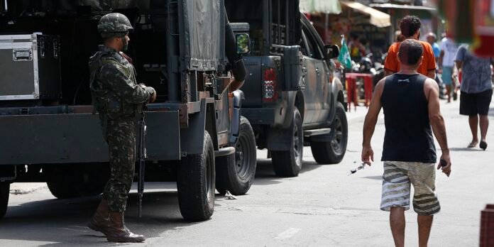 Soldier in camouflage with a rifle standing beside a large armored vehicle on a busy urban street.