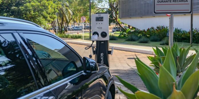 Electric car plugged into a public charging station with a no-parking sign in the background and greenery in the foreground viewed at curbside.