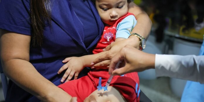 Child wearing red outfit sits on a lap while a healthcare professional administers a injection into the child’s arm.