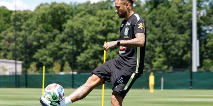 Male soccer player in a black training kit kicks a ball during drills on a sunny training field, with yellow poles and a green fence in the background.