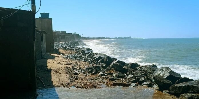 Row of dark buildings lining a sandy beach with a rocky breakwater extending into the sea under a clear blue sky.