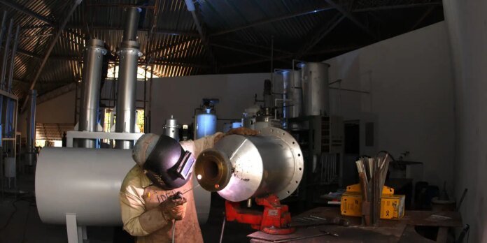Welder in protective gear working on a metal flange clamped in a red vise inside an industrial workshop with large pipes and equipment nearby.