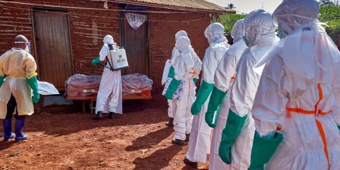 Healthcare workers in full PPE stand in a line near a covered coffin outside a mud-brick building, preparing for a burial.