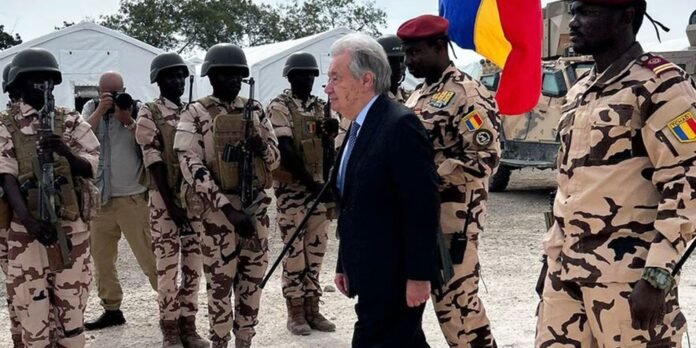 A man in a dark suit walks between a line of soldiers in desert camouflage uniforms with helmets and rifles, near tents at a military base.
