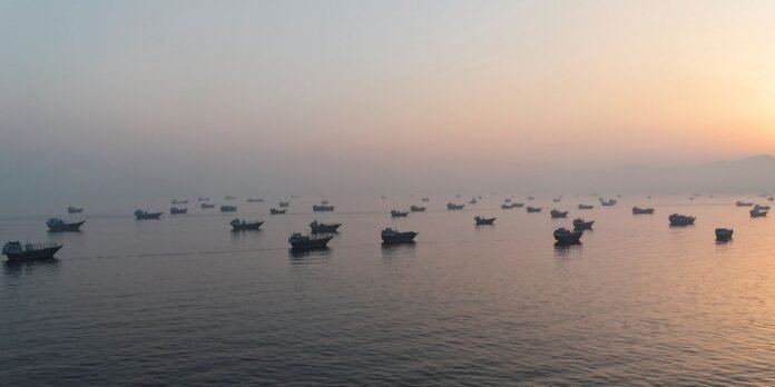 Many small boats dot a calm sea at sunset, with a hazy horizon and distant mountains bathed in warm light to the right edge of the image.