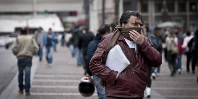 Man in a burgundy jacket covers his mouth with a scarf while holding a stack of papers on a busy city sidewalk.