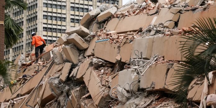 Construction workers in orange safety gear on a collapsed concrete building site filled with rubble and rebar in an urban area.