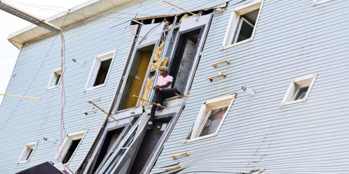 Construction worker sits on a metal platform at a damaged doorway of a blue apartment building, with exposed insulation and framing visible around the opening.