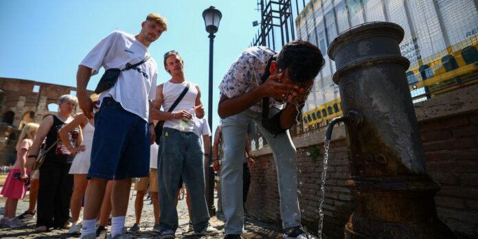 Group of young tourists gather around a public drinking fountain on a sunny street; one man drinks while others wait nearby.