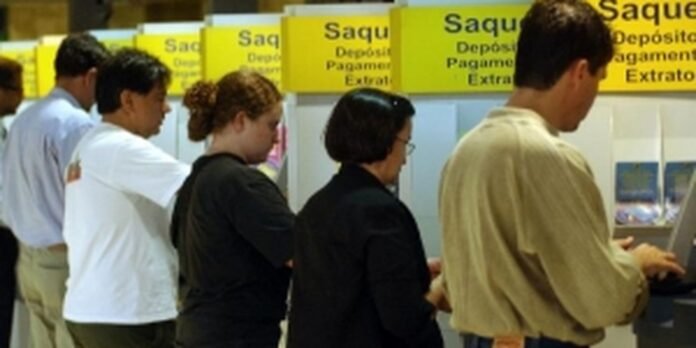 People stand in line at bank counters beneath yellow signs advertising withdrawals, deposits, and statements.