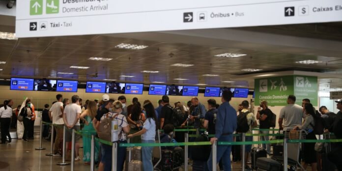 Crowded airport arrivals hall with travelers lined up at check-in counters beneath overhead signs showing Domestic Arrival and taxi icons.