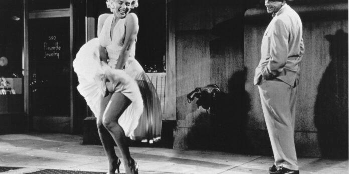 Woman in a white sleeveless dress dances on a sidewalk as a man in a light suit watches from the right, storefront in the background.