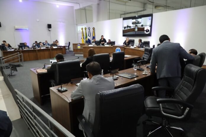 Council chamber scene with officials seated around a U-shaped conference table, flags behind the dais, and a large display screen showing a video conference.