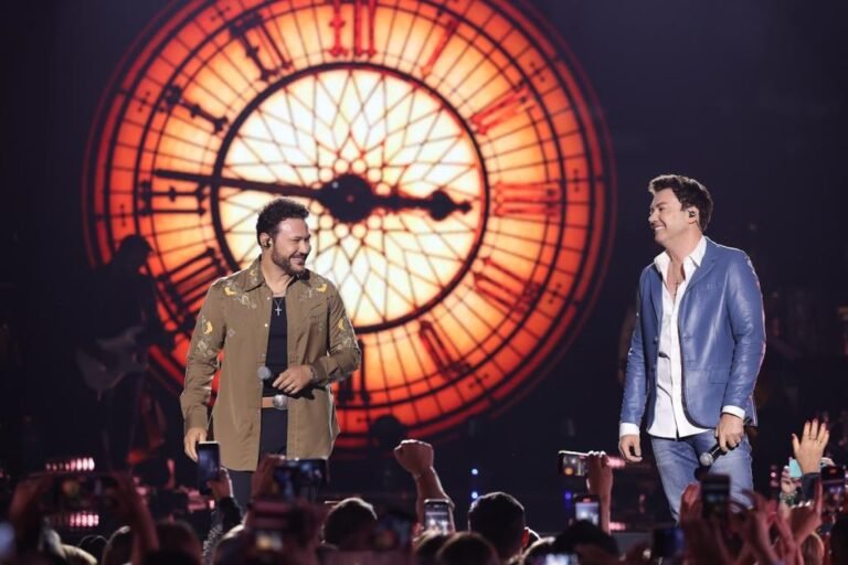 Two male performers on a stage with a giant illuminated clock backdrop, microphones in hand, audience cheering in the foreground.