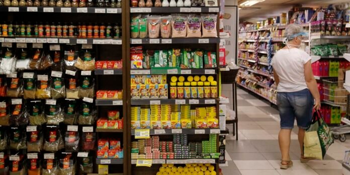Woman in a white shirt, denim shorts, mask and hairnet stands with a reusable bag in a grocery store aisle, facing away from the camera, with shelves stocked with spices and packaged foods on both sides bathed in store light.