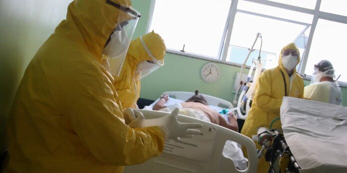 Medical team in yellow hazmat suits and masks attend a patient in a hospital bed with ventilator equipment nearby.