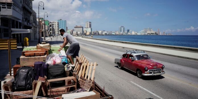 Man arranging luggage and furniture on a roadside cart while a vintage red car drives by along a waterfront avenue with city buildings in the background.