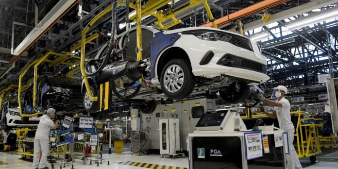White car on an assembly line suspended by robotic equipment in a factory setting, with workers in white uniforms assembling components.