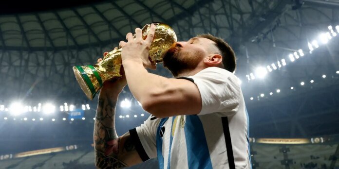 Soccer player in a white and blue jersey kissing a gold trophy on a bright stadium stage after a victory.