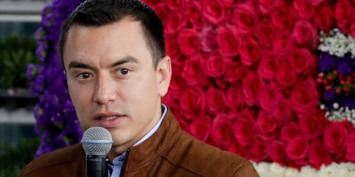 Man in a brown jacket speaks into a handheld microphone against a backdrop of pink roses and purple flowers.