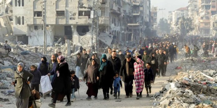 A large group of civilians walking through a rubble-strewn street in a war-torn city, carrying belongings and children.