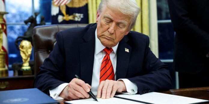 Man in a navy suit with a red striped tie signs a document at a desk in a formal office, American flag pin visible on lapel.