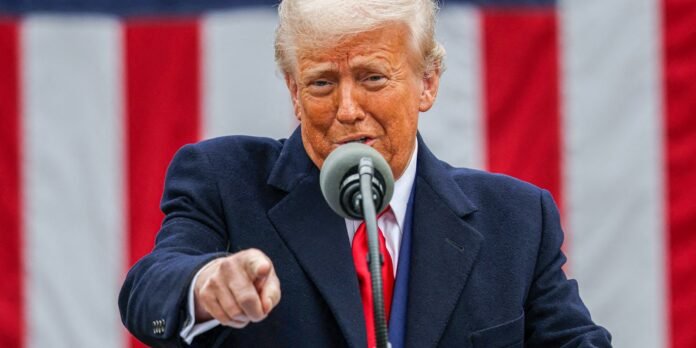 Politician at a podium, pointing toward the audience, with a red tie and dark coat, against a red-white-blue striped backdrop.