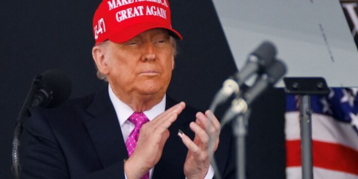 Man in a dark suit and bright pink tie wearing a red 'Make America Great Again' cap, standing by microphones at a podium, hands clasped slightly apart.