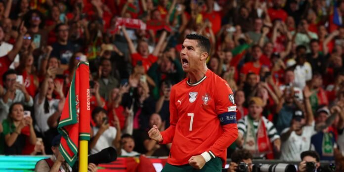 Portuguese footballer in a red kit celebrating a goal as fans cheer in the stands with red shirts nearby.
