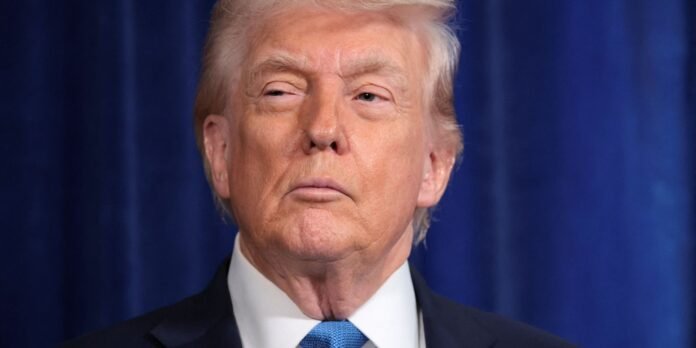 Close-up portrait of a light-haired man in a dark suit and blue tie against a blue curtain backdrop.