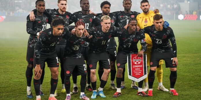 Canada national men's soccer team posing for a group photo on the pitch before a Canada–Tunisia match, with a pennant held by players in black kits on a grassy field.