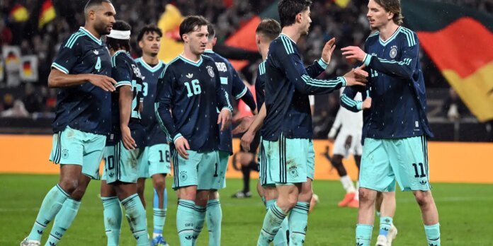 German national team players celebrate on the field, high-fiving after a match in navy and light-blue kit under stadium lights, rain visible in the air