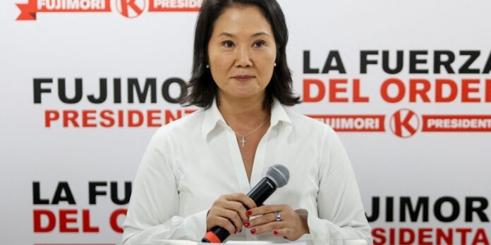 Woman in a white blouse speaking at a press conference, holding a microphone, with bold red/black campaign signage behind her.