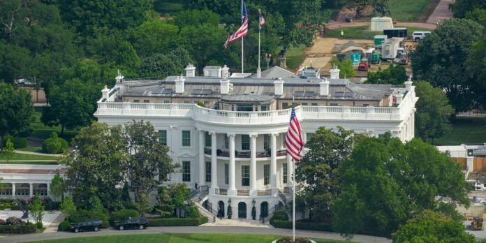 White neoclassical mansion with curved portico and tall columns, two American flags flying nearby amidst green trees.