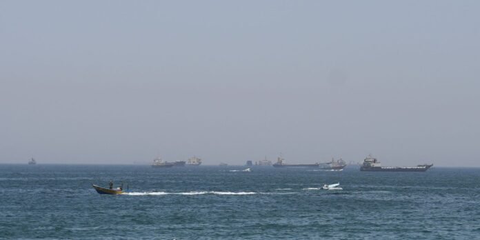 Small speedboat with two people in the foreground speeds across blue water, with a distant fleet of ships on a hazy horizon.