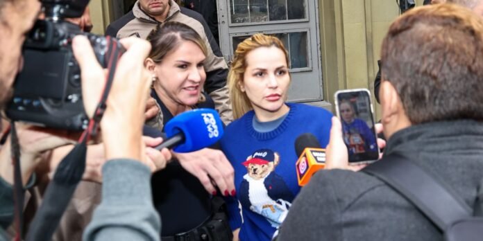 Woman in a blue sweater being interviewed by reporters amid a press scrum outside a building, with cameras and microphones nearby.