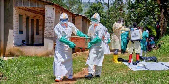 Medical responders in full protective suits carry a wooden coffin outdoors, with others in PPE nearby at a rural facility site.