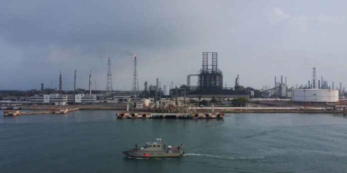 Industrial port scene with a small tugboat on calm water and large refinery structures in the background.